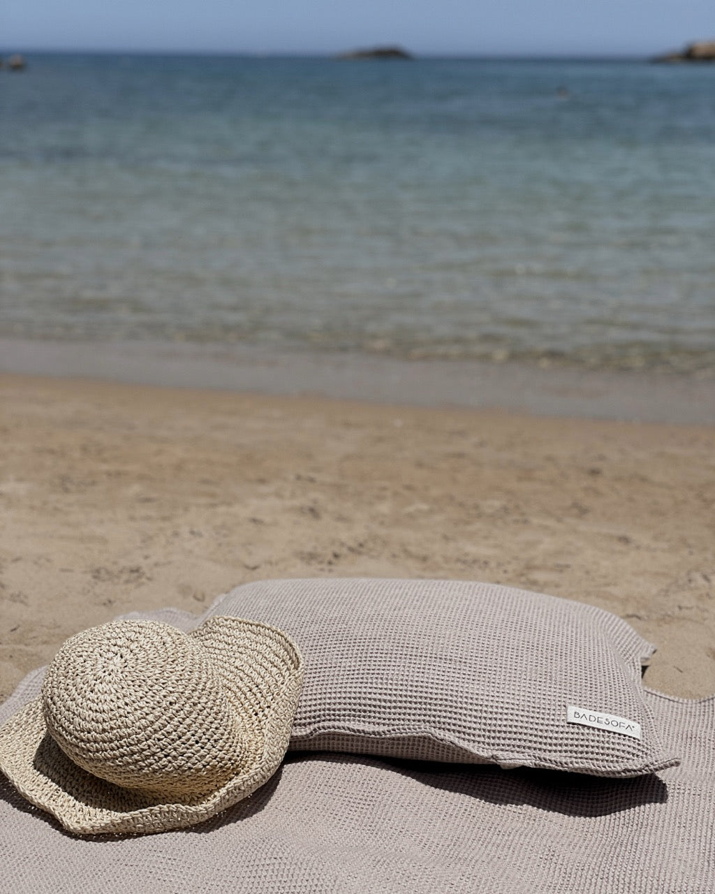 Nahaufnahme eines Strandtuchs und Kopfkissen. Ein Strohhut liegt dekorativ mit auf dem Strandtuch, im Hintergrund das Meer.