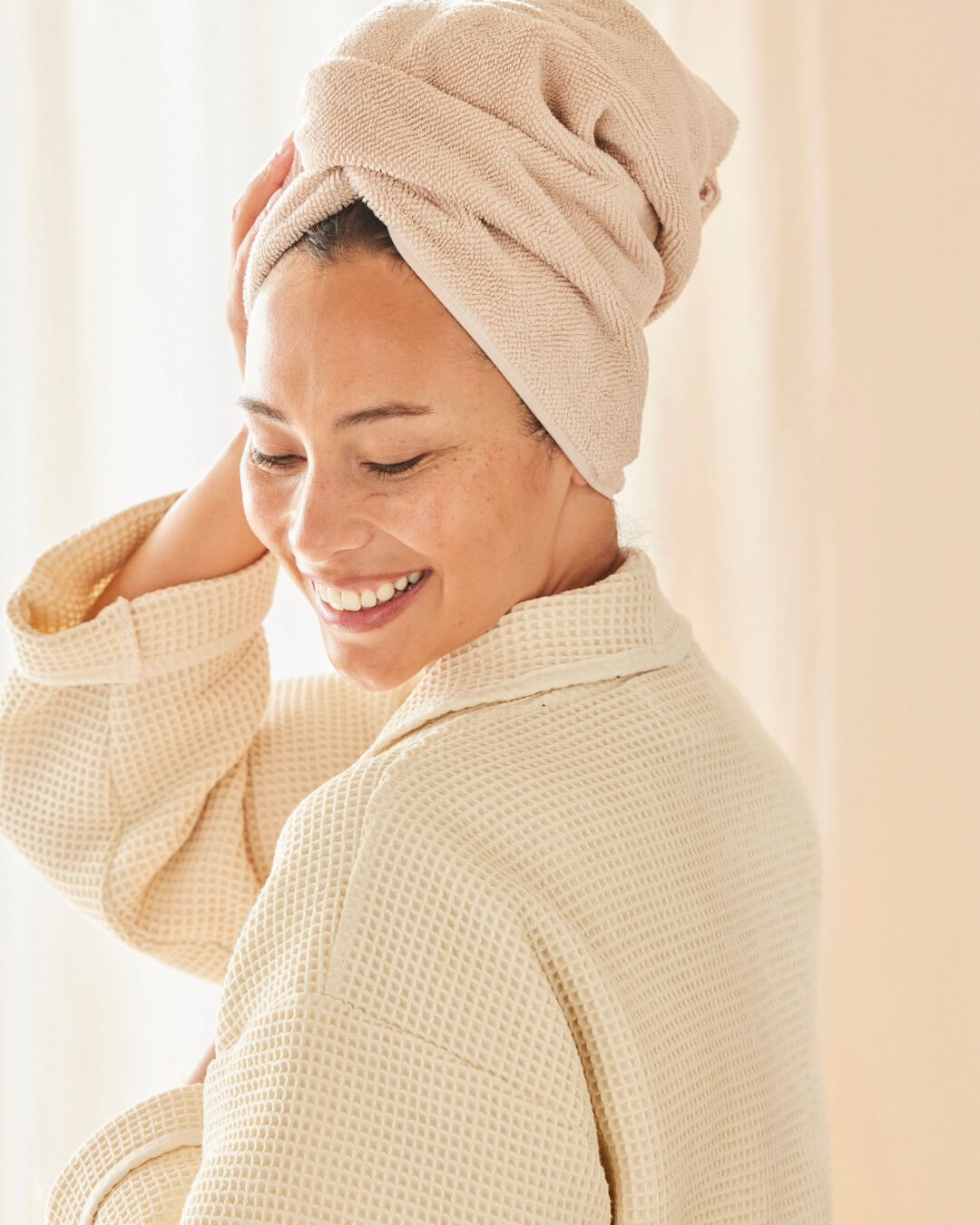 Frau steht im Bademantel (Farbe Nature) im Bad und that die Haare in ein Handtuch gewickelt. 