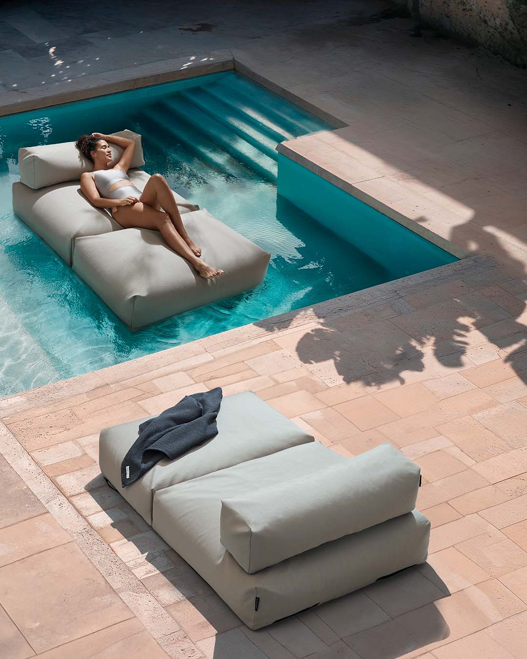 Eine Frau liegt entspannt auf einem Poolsofa double in Beige, das im Pool schwimmt. Am Rand des Pools, auf der Terrasse steht ein weiteres Poolsofa. $ Poolsofa Double | Floating Lounger | Beige