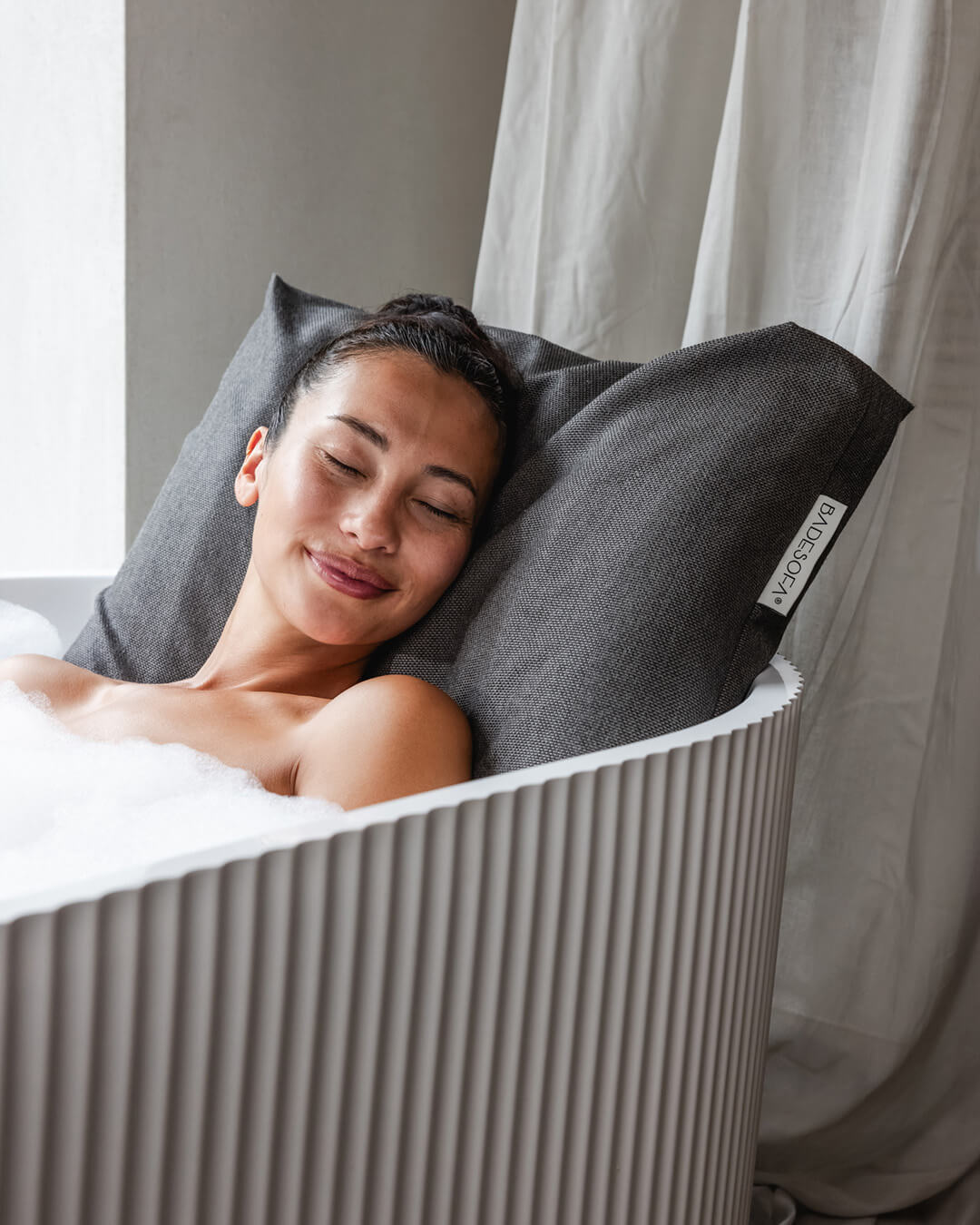 Mocha Badesofa Rückenkissen (Size Large) in einer freistehenden Badewanne, mit lächelnder Person, die sich entspannt anlehnt. 