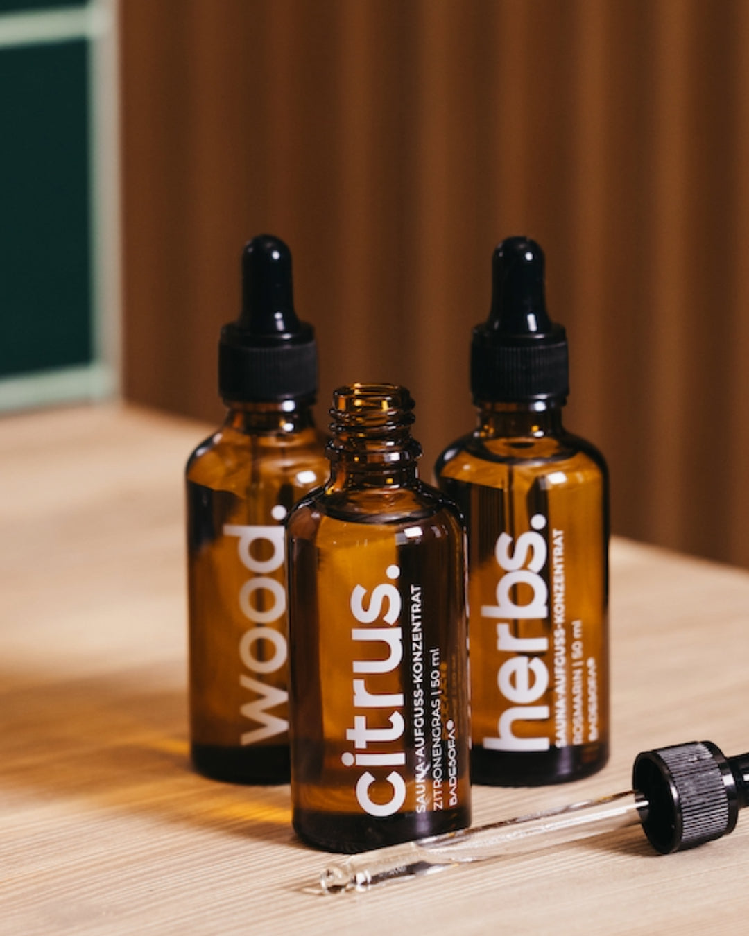 Nahaufnahme von drei kleinen Glasfläschchen mit Pipette, die drei Duftrichtungen Saunaaufguss-Konzentrat enthalten. Die Duftrichtungen sind Wood, Cirtus und Herbs. Die Pipette einer Flasche liegt vor den Fläschchen.