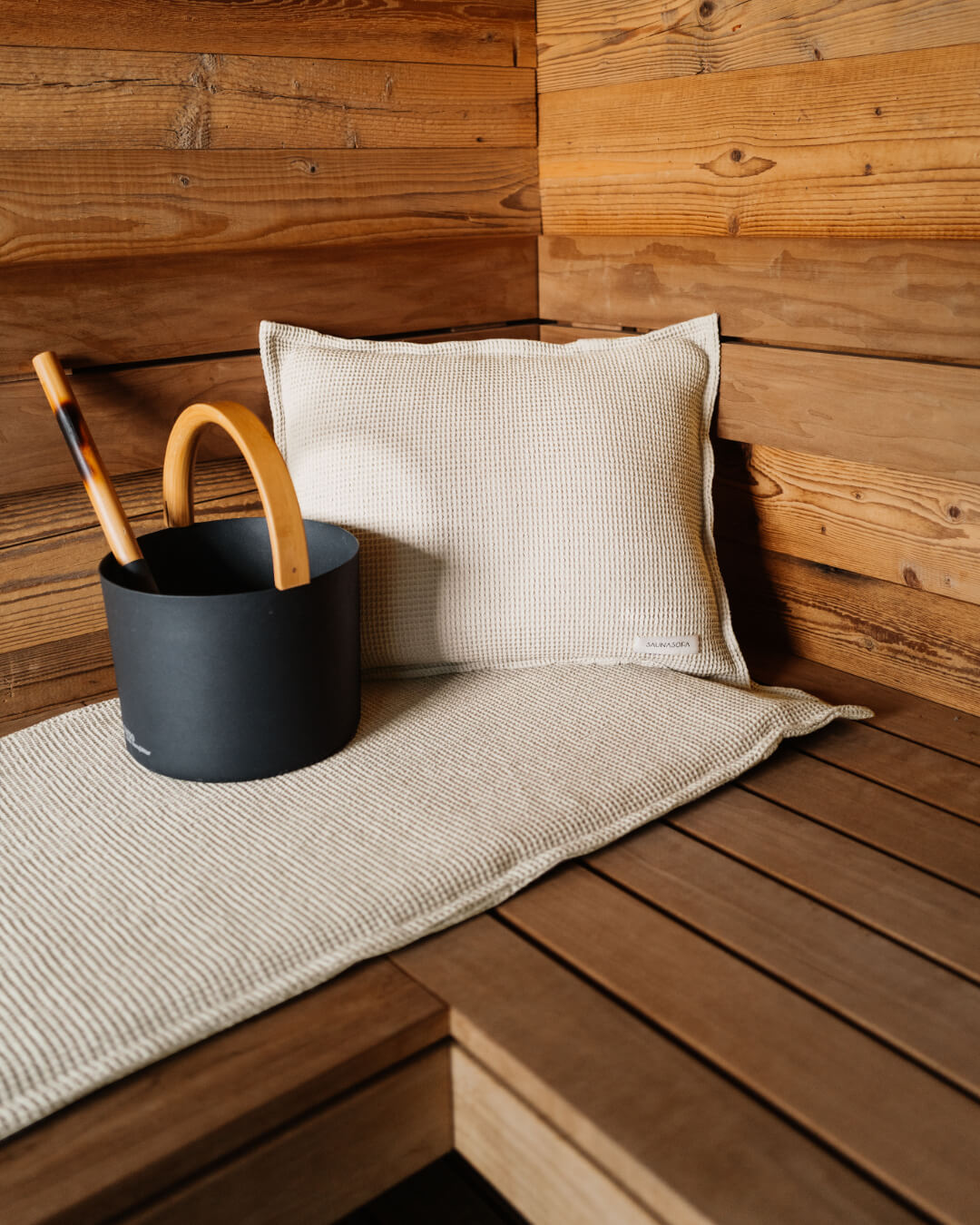 Nahaufnahme von einem Saunakopfkissen und einer Saunaauflage in der Farbe Nature. Daneben steht der schwarze Badesofa Saunakübel mit Kelle für Aufgüsse. 