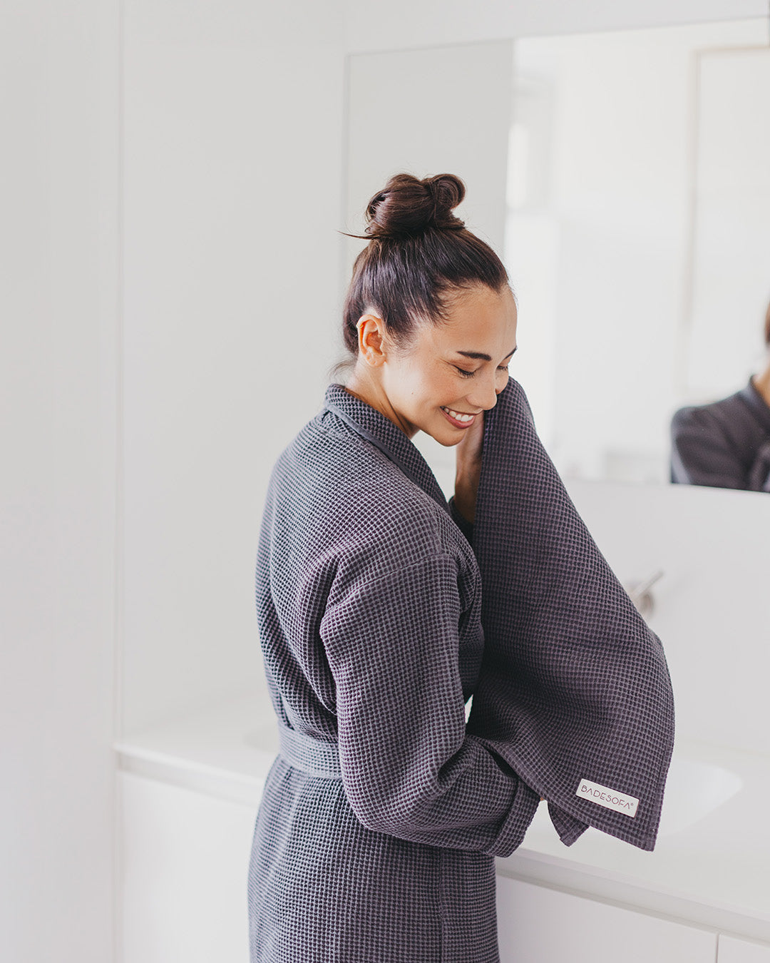 Frau steht im Bademantel (Farbe Anthrazit) vor dem Spiegel und trocknet sich das Gesicht ab. 